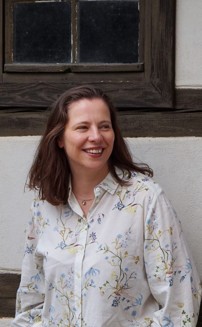 Diane Maren Jende mit langen, braunen Haaren, lächelt vor einer grünen, bewachsenen Kulisse.